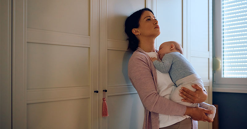 Parent leaning against a wall, gazing at ceiling while holding infant.