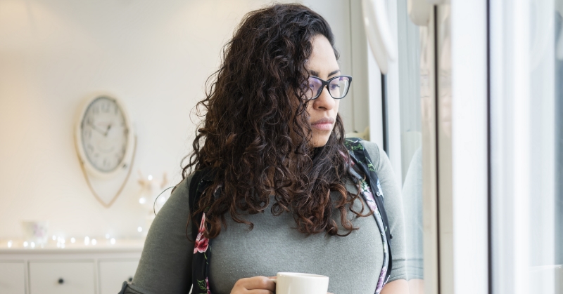 Pregnant person holding a cup, looking out a window.