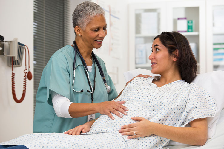 A pregnant woman sits up on a hospital bed and is speaking with a health care provider.