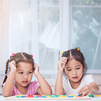 Two children seated at table. One inserts a piece to a puzzle while the other watches.