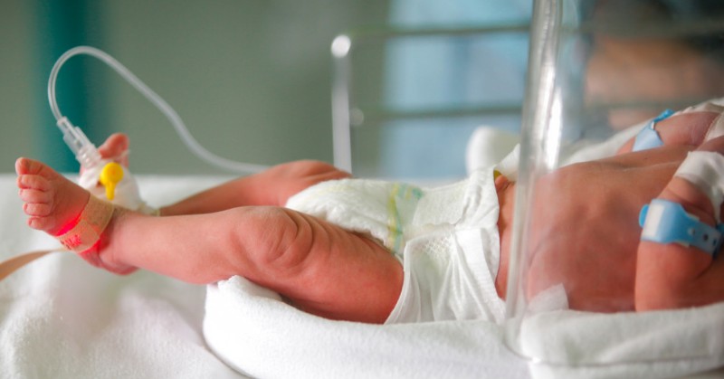 An infant lying in an incubator. The infant is wearing a diaper, two medical bracelets, and a foot monitor.