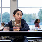 Child seated, alone, at a table, staring into the distance. Other children seated in the far background.