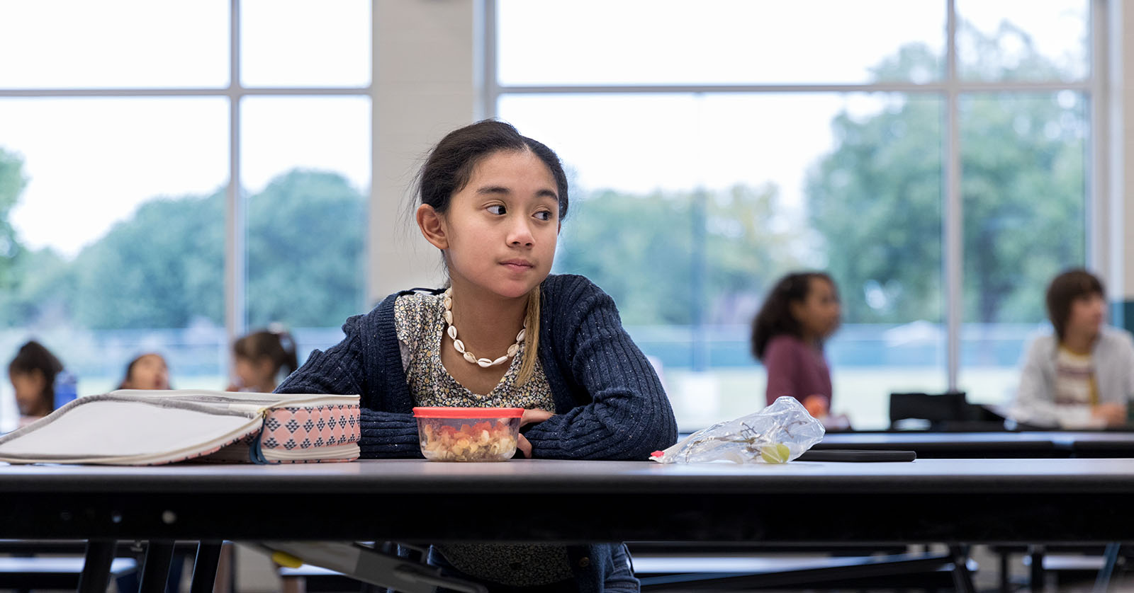 Child seated, alone, at a table, staring into the distance. Other children seated in the far background.