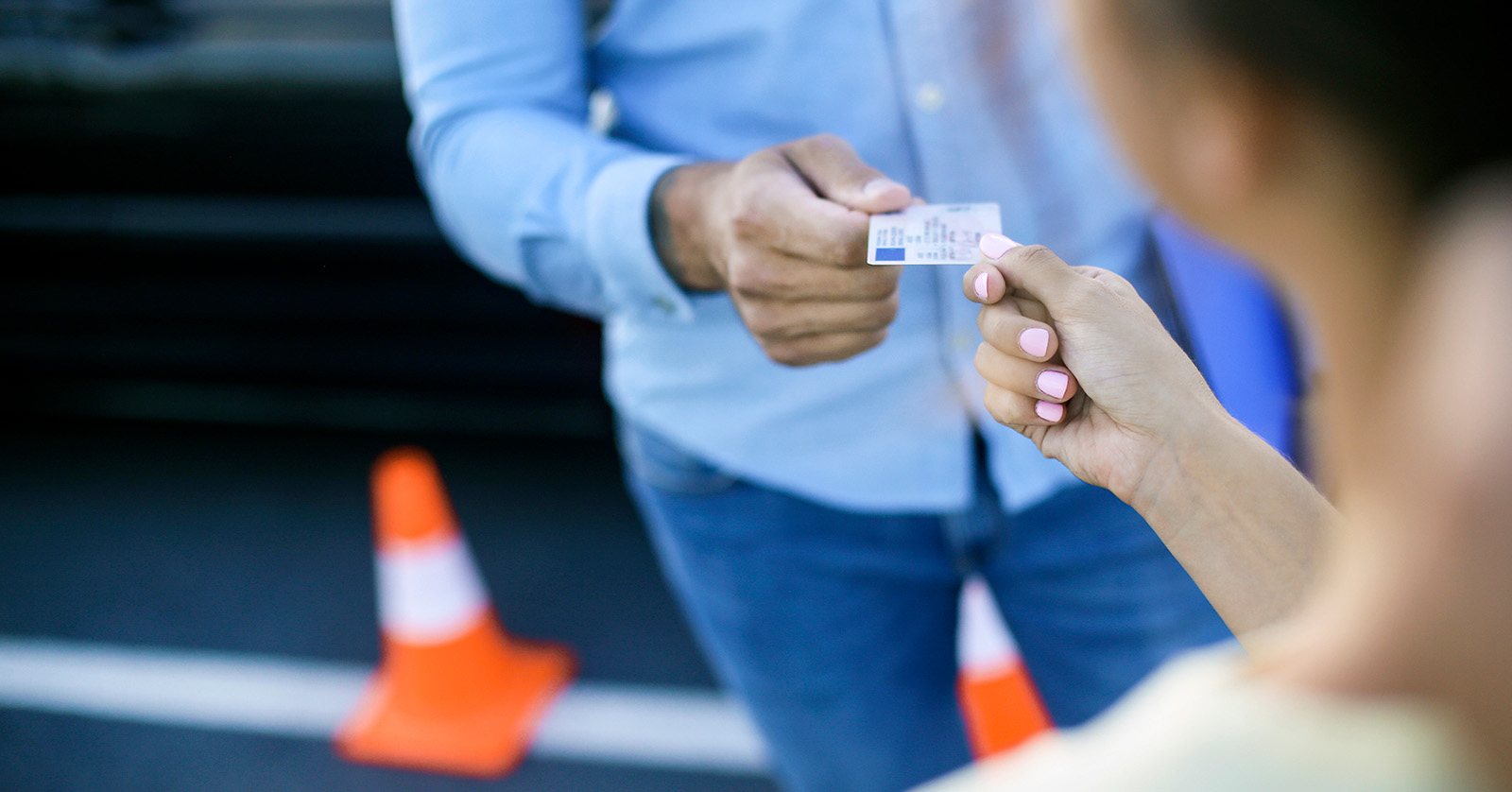 Close-up view of one person handing a driver's license to another person. Two orange traffic cones are on the ground in the background.