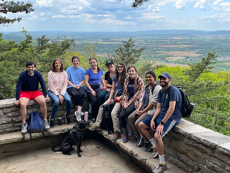 Nine smiling people sit on a stone wall with greenery, hills, and a blue sky in the background. Two black dogs sit at the feet of the the people.