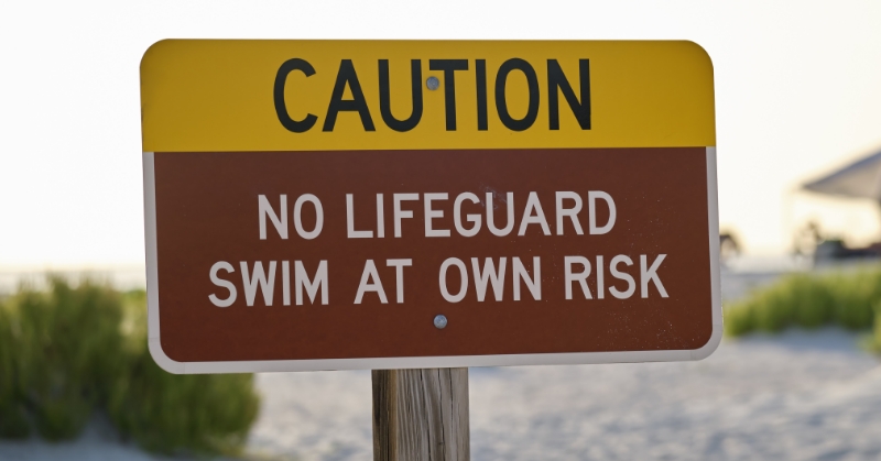 A sign reading “Caution, no lifeguard, swim at own risk” appears in the foreground. A sandy beach with dune grasses appears blurred in the background.