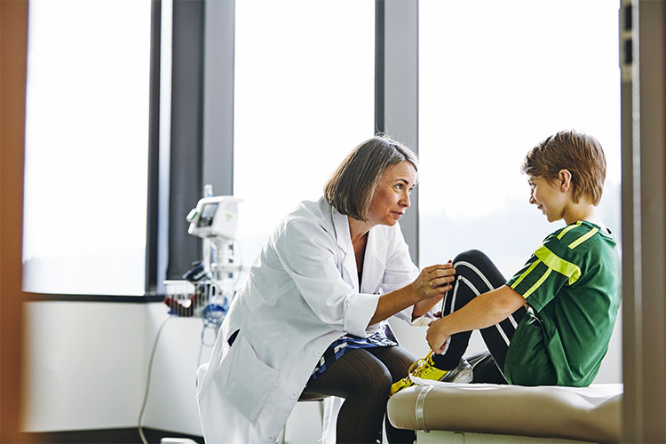 A young person sits with their arms around their bent knees, in front of a health care provider.