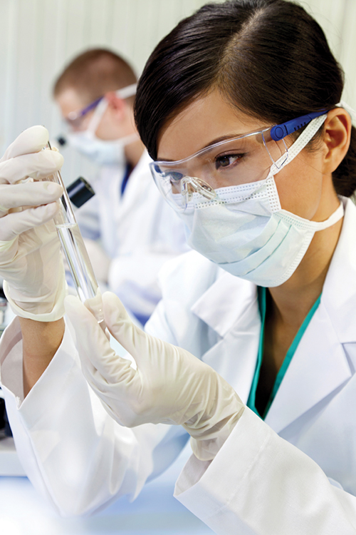 Researcher wearing safety goggles, surgical mask, and latex gloves, examining the contents of a test tube.