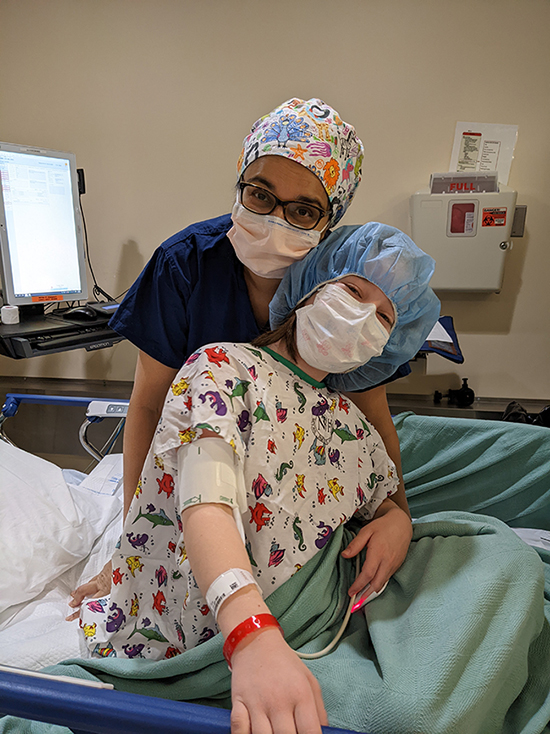 A female doctor wearing a hair covering, glasses, and a mask hugs a girl from behind. The girl is sitting on a hospital bed with a blood pressure cuff around her arm and wearing a hair net, mask, and hospital gown.