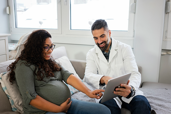 A pregnant woman seated on a sofa with a health care provider, discussing information on a tablet computer.