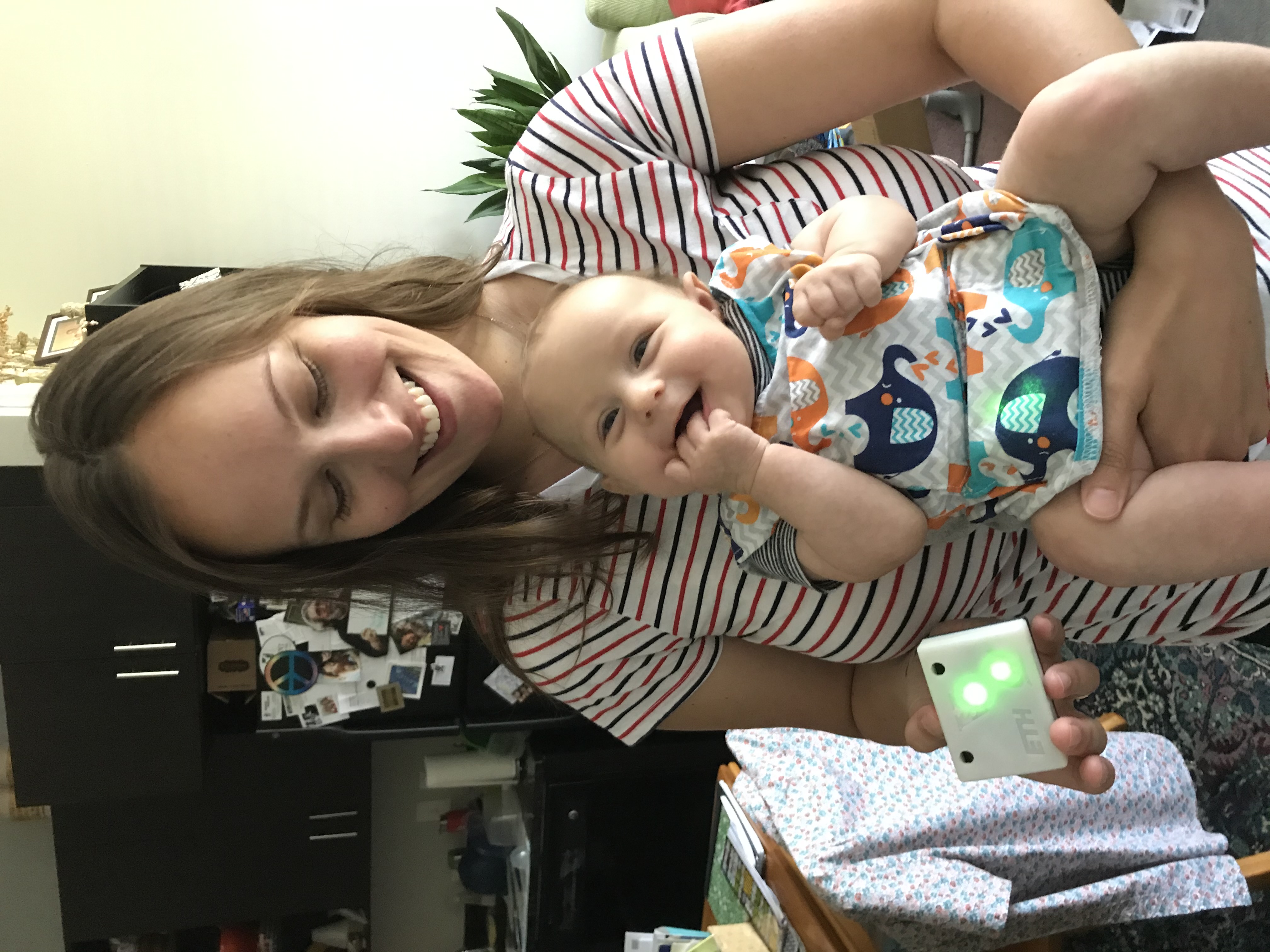 A smiling woman in a striped shirt holds a baby wearing an elephant bib in her left arm. Her right hand holds a small rectangular device with two glowing green dots.