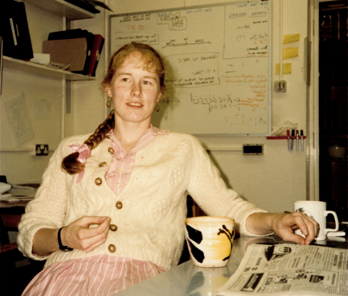 Dr. Dasso is sitting at a table with coffee mugs and a newspaper. Behind her is a white board and shelving.