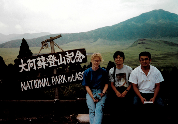 The group is sitting next to a wooden sign that has white letters. The top of the sign has Japanese writing and the bottom says, “NATIONAL PARK mt ASO.” The mountain is visible in the background.