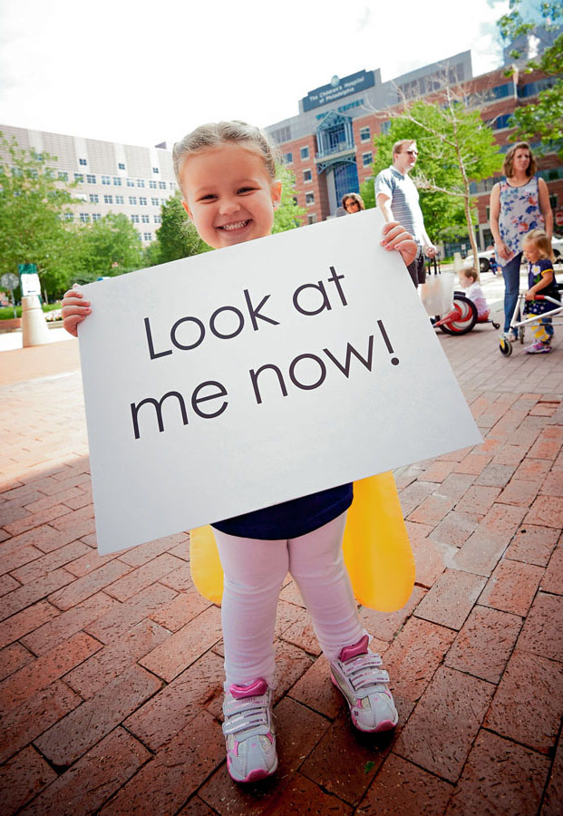 A young girl standing on a brick walk holding a sign that reads, “Look at me now!”